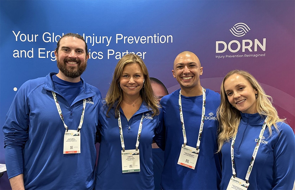 DORN team members smiling and standing together at a conference booth, wearing branded blue apparel, representing the company’s commitment to injury prevention and workplace ergonomics.